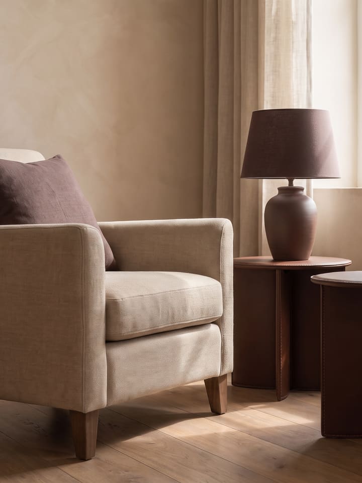 Detalhe do mesmo ambiente: poltrona de linho com almofada cacau, mesas laterais de couro cacau e luminária junto à janela com cortina de linho.